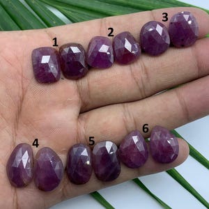 May include: A collection of six faceted, deep red gemstones. The stones vary in shape, including rectangular, oval, and teardrop. Each gem has a polished, reflective surface, showcasing the internal facets and color variations.