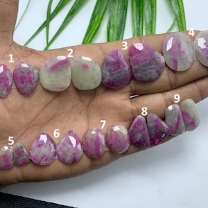 May include: A collection of nine faceted gemstones, primarily in shades of pink and white, are displayed. The stones vary in shape, including oval and teardrop forms. The gemstones are arranged on a light-colored surface, showcasing their polished surfaces and internal patterns.