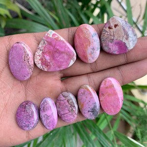 May include: A collection of polished gemstones in various shapes, including oval and triangular. The stones display a range of pink and magenta hues, with some featuring white and brown veining. The gemstones are displayed on a hand.
