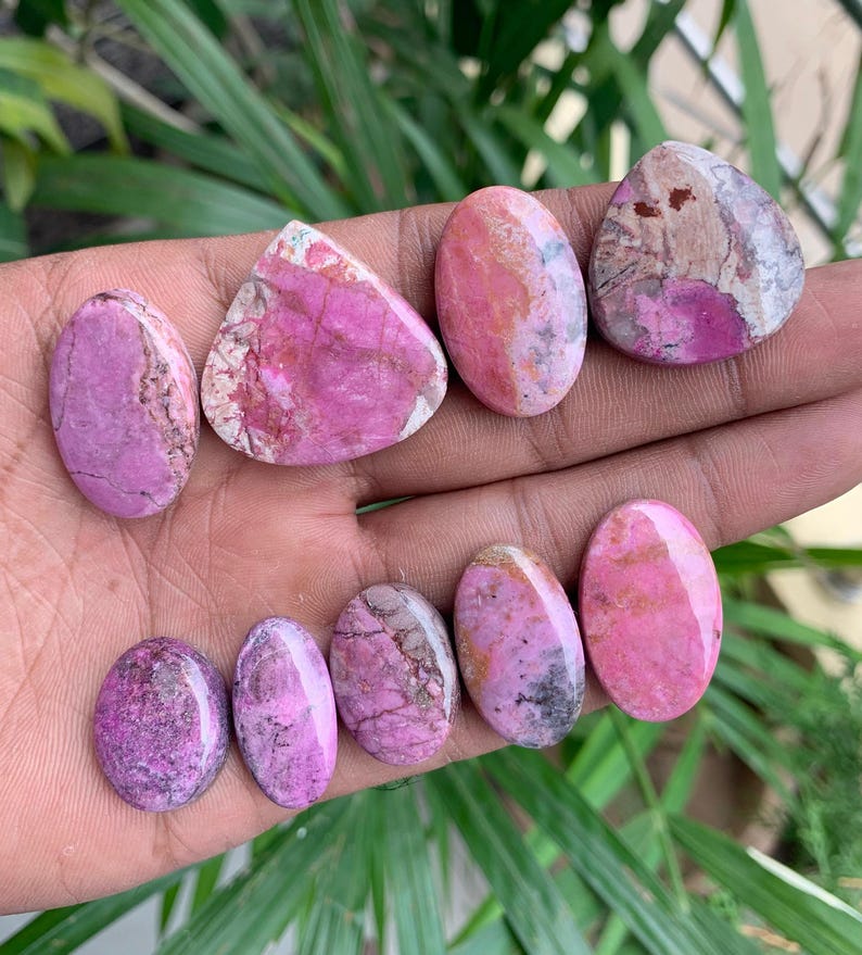 May include: A collection of polished gemstones in various shapes, including oval and triangular. The stones display a range of pink and magenta hues, with some featuring white and brown veining. The gemstones are displayed on a hand.