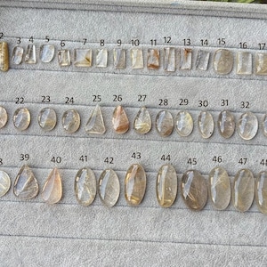 Op de afbeelding: Een verzameling van 50 genummerde, gepolijste edelstenen, tentoongesteld op een grijze tray. De stenen variëren in vorm, waaronder rechthoeken, ovalen en druppels. De edelstenen zijn voornamelijk helder met gouden insluitsels.