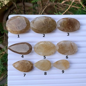 May include: Nine polished, golden-brown gemstone cabochons with internal inclusions are arranged on a white surface. The stones vary in shape, including oval and teardrop forms. The gems have a translucent quality, with visible internal structures.
