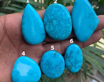 May include: Six polished turquoise gemstones in various shapes are displayed. The stones range in color from light to medium blue, with some showing dark veining. The shapes include teardrop, oval, and rounded forms. The gemstones are held in a hand.