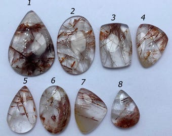 May include: Eight polished gemstones in various shapes, including teardrop, oval, and rectangular. The stones are clear with reddish-brown inclusions, creating unique patterns within each piece. The gemstones are displayed on a light-colored surface.