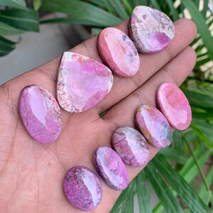 May include: A collection of polished gemstones in shades of pink and purple, held in a hand. The stones vary in shape, including oval and teardrop, with unique patterns and textures. The gemstones are displayed against a backdrop of green foliage.