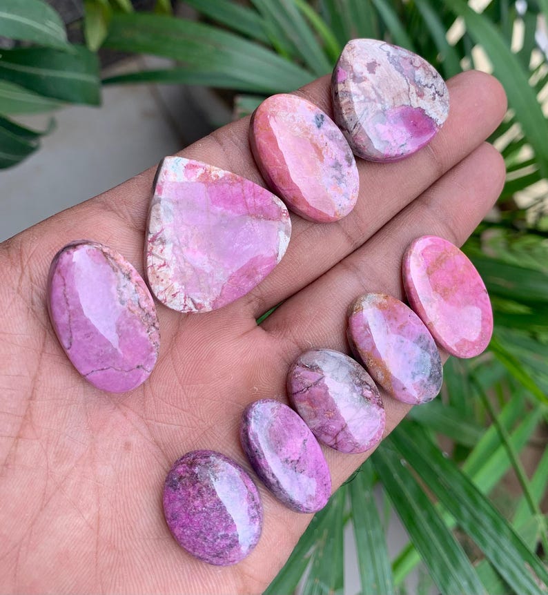 May include: A collection of polished gemstones in shades of pink and purple, held in a hand. The stones vary in shape, including oval and teardrop, with unique patterns and textures. The gemstones are displayed against a backdrop of green foliage.