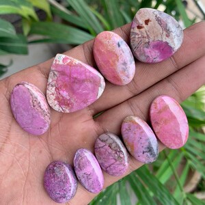 May include: A collection of polished gemstones in shades of pink and purple, with unique patterns and textures. The stones vary in shape, including oval and irregular forms. The gemstones are displayed against a natural background.