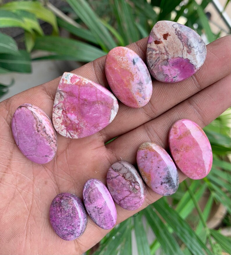 May include: A collection of polished gemstones in shades of pink and purple, with unique patterns and textures. The stones vary in shape, including oval and irregular forms. The gemstones are displayed against a natural background.