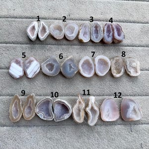 May include: A collection of twelve polished geode specimens, each with a unique shape and colour. The geodes are open-faced, revealing crystal formations in shades of white, grey, purple, and brown. The sizes vary across the collection.