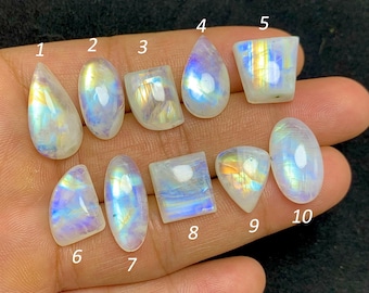 May include: Ten iridescent moonstone gemstones in various shapes, including teardrop, oval, and rectangular. The stones display a range of colors, including blue, yellow, and white, and are held in a hand against a dark background.