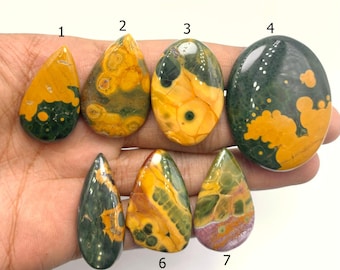 May include: Seven polished gemstones in various shapes, including teardrop and oval. The stones feature a mix of colors, including yellow, green, and brown. The gemstones are displayed against a light background.