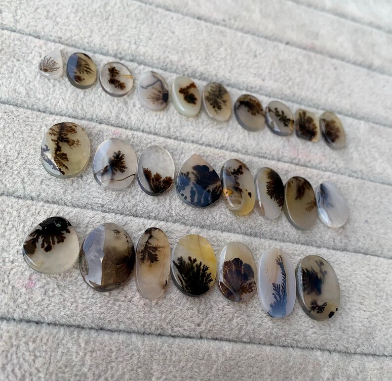 May include: A collection of polished dendritic agate gemstones in various oval and teardrop shapes. The stones display intricate, tree-like patterns in shades of brown and black against a translucent, creamy background. The gemstones are arranged on a gray surface.