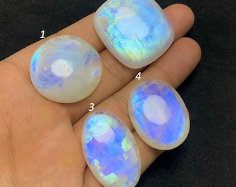 May include: Four polished moonstone gemstones in various shapes: round, square, and oval. The stones display a milky white base with iridescent blue and yellow flashes. The gemstones are held in a hand against a dark background.
