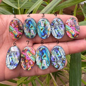 May include: Nine oval-shaped abalone shell cabochons, each displaying iridescent colors of blue, green, pink, and purple. The cabochons are arranged in two rows, showcasing the natural patterns and textures of the shells. These are ideal for jewelry making.