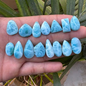 May include: A collection of polished, light blue larimar gemstones in various shapes, including teardrops, ovals, and rectangles. The stones display a marbled pattern, with white veining throughout. The gemstones are displayed on a hand.