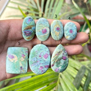 May include: A collection of seven polished gemstones, each with a unique shape. The stones feature a mix of green, blue, and pink hues. The gemstones are oval, rectangular, and teardrop-shaped, showcasing natural patterns and textures.