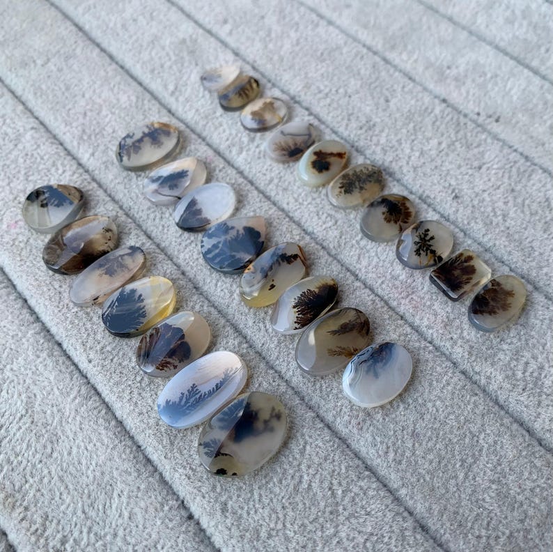 May include: A collection of polished dendritic agate gemstones in various oval and rectangular shapes. The stones display natural patterns in shades of brown, black, and white, resembling miniature landscapes. The gemstones are arranged on a gray, textured surface.
