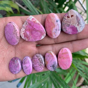 May include: A collection of polished gemstones in various shapes, including oval and triangular. The stones display a range of pink and purple hues with unique veining and patterns. The gemstones are held in a hand.