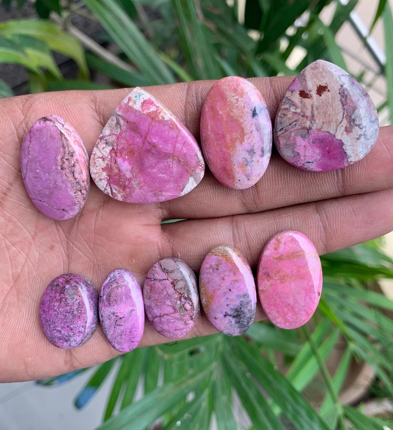 May include: A collection of polished gemstones in various shapes, including oval and triangular. The stones display a range of pink and purple hues with unique veining and patterns. The gemstones are held in a hand.