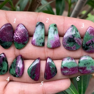 Op de afbeelding: Tien groene en roze edelsteen cabochons, genummerd van 1 tot en met 6, gerangschikt in rijen op een hand. De edelstenen zijn in verschillende tinten groen en roze, sommige met zwarte insluitsels.
