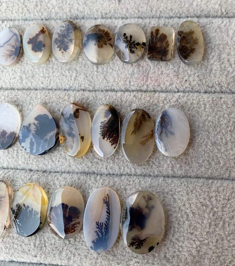 May include: A collection of polished dendritic agate gemstones in various oval and rectangular shapes. The stones display intricate, tree-like patterns in shades of brown and black against a translucent, milky background. The gemstones are arranged on a gray surface.