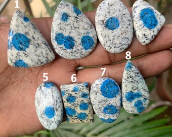 May include: A collection of eight K2 stone cabochons, each with a unique shape. The stones feature a speckled white and gray base with vibrant blue spots and patterns. The cabochons are displayed against a natural background.