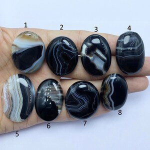 May include: Eight black and white agate cabochons with different patterns. The cabochons are oval shaped and have a smooth, polished finish.
