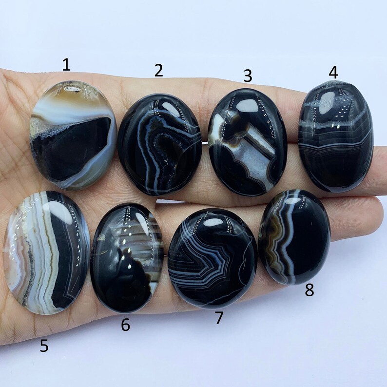 May include: Eight black and white agate cabochons with different patterns. The cabochons are oval shaped and have a smooth, polished finish.