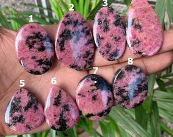 Könnte beinhalten: Acht polierte Rhodochrosit-Edelsteine in verschiedenen Formen, darunter oval und tropfenförmig. Die Steine zeigen einen leuchtenden rosa Farbton mit auffälligen schwarzen Aderungsmustern. Die Edelsteine werden in einer Hand gehalten, mit grünem Laub im Hintergrund.