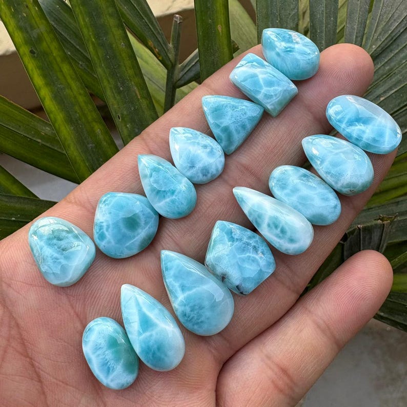 May include: A collection of polished larimar gemstones in various shapes, including teardrop, oval, and rectangular. The stones display a light blue color with white marbling, resembling ocean waves. The gemstones are held in a hand, with green leaves in the background.