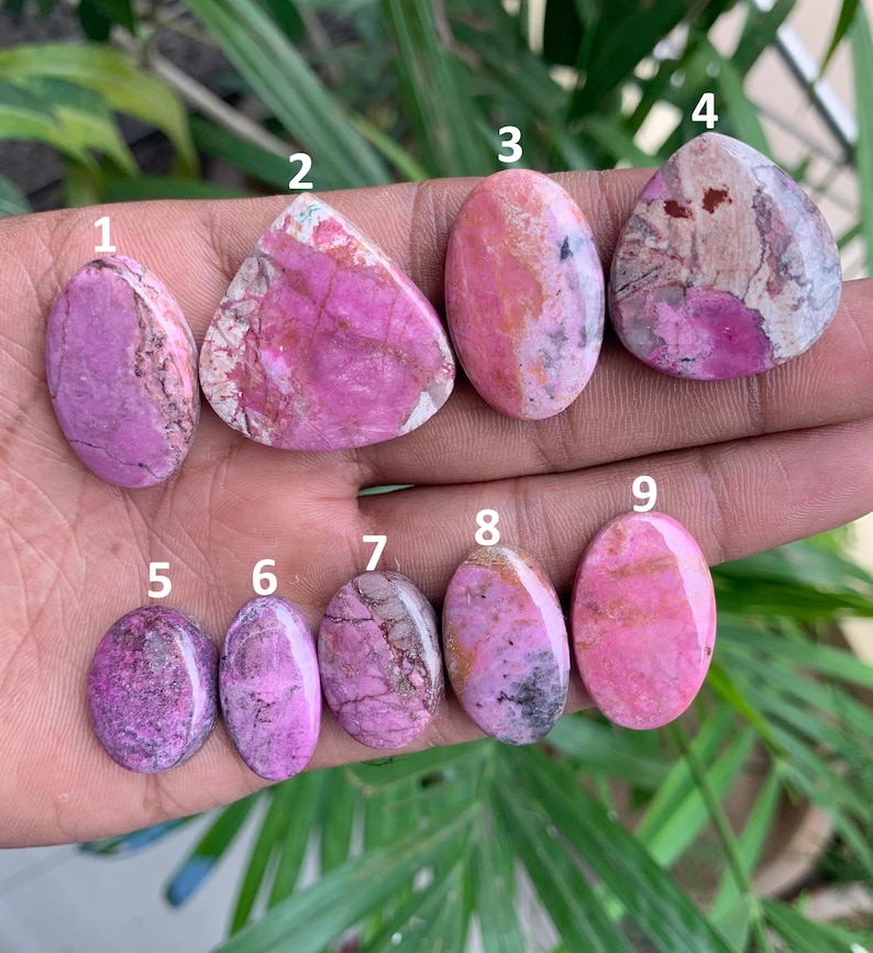 May include: A collection of nine polished gemstones in various shapes, held in a hand. The stones display shades of pink, purple, and white, with unique veining and patterns. The gemstones are oval, triangular, and rounded.