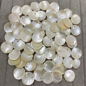 May include: A pile of iridescent, round, white mother-of-pearl shell pendants. Each pendant has a small hole for threading. The pendants are scattered on a grey surface, creating a natural, organic aesthetic.