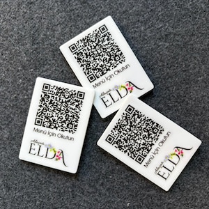 Puede incluir: Tres tarjetas rectangulares blancas con códigos QR en blanco y negro. Las tarjetas tienen el texto "Menü için Okutun" y "Alaçatı ELDA" con detalles florales. Las tarjetas están sobre una superficie gris.