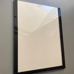 May include: A rectangular, white framed picture with a black border. The frame is mounted on a gray wall. The picture's surface reflects light, showing a glimpse of the outside environment.