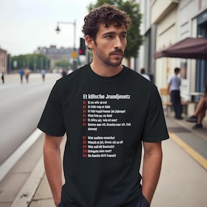 Peut inclure: T-shirt noir avec texte blanc en allemand. Le texte dit "Et kölsche Jrundjesetz" et présente une liste de dictons. Le t-shirt est porté par une personne dans une rue de la ville.