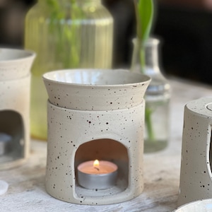 以下が含まれることがあります： 白い斑点模様のセラミック製アロマディフューザー。小さなティーライトキャンドルが入っています。ディフューザーの上部と下部に丸い開口部があります。ディフューザーは白い表面の上に置かれています。