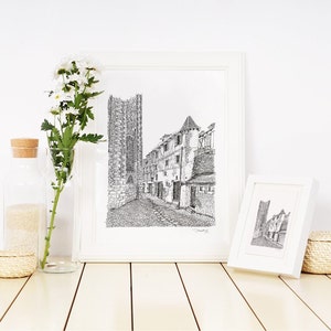 May include: Two black and white ink drawings of a street scene with buildings and a church tower in white frames on a white wood surface with a vase of white flowers and a glass jar with sand.