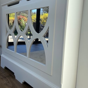 May include: A white radiator cover featuring a rectangular frame with a mirrored panel insert. The mirrored section displays a decorative cutout pattern consisting of four interconnected arch shapes. The structure has a flat top surface and a base.