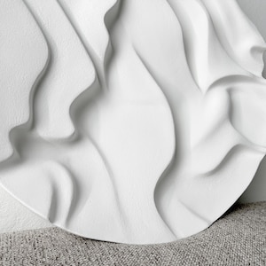 May include: White circular wall sculpture with a textured, wavy design. The sculpture has a 3D effect, creating a sense of depth and movement.