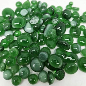 May include: A selection of green glass pebbles in assorted shapes and sizes. The pebbles vary in shades of green, from light to dark, with some appearing milky. The smooth, rounded forms reflect light, producing a glossy finish.