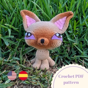 Puede incluir: Una muñeca de zorro de ganchillo con detalles marrones, beige y rosas, sobre un fondo de hierba verde. El zorro tiene orejas grandes, detalles de ojos morados y una nariz negra. La imagen incluye el texto "Crochet PDF pattern".