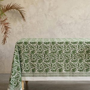 May include: A green and white floral tablecloth. The tablecloth is draped over a wooden table. The floral pattern is a repeating design of leaves and flowers. The tablecloth has a white border with a green floral design.