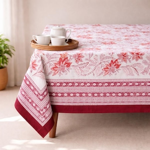 May include: A white tablecloth featuring a floral design in red and pink hues. The tablecloth has a decorative border and a deep red edge. A wooden tray with white teacups and a teapot rests on the table.