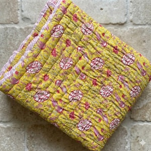 Puede incluir: Edredón floral amarillo doblado con un patrón repetido de grandes flores blancas y rosas. El edredón tiene una textura acolchada y un borde a rayas rosa y blanco. El edredón está doblado y apilado.