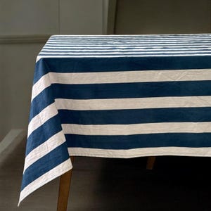 Puede incluir: Un mantel rectangular con rayas anchas alternas en azul marino y blanco. El mantel está colocado sobre una mesa de madera, con las rayas corriendo horizontalmente por la parte superior y verticalmente por los lados.