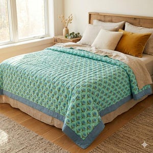 Op de afbeelding: Een turquoise en blauw bloemenpatroon quilt op een bed. De quilt heeft een blauwe rand en wordt gecombineerd met beige en gele kussens. Het bed is van hout en staat op een vloerkleed.