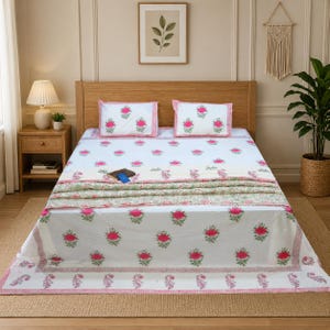 Puede incluir: Una colcha floral blanca con fundas de almohada a juego. La ropa de cama presenta un patrón repetido de rosas rosas y hojas verdes. Los bordes están adornados con un borde rosa. Una colcha doblada con un estampado floral está al pie de la cama.