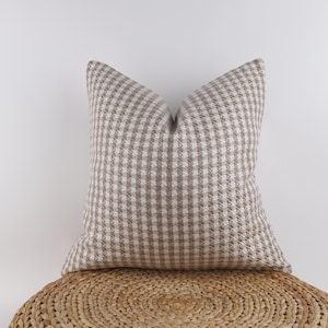 May include: A square decorative pillow with a brown and white checkered pattern. The pillow is made of a textured fabric and is resting on a woven, circular surface. The pillow has a neutral color palette.