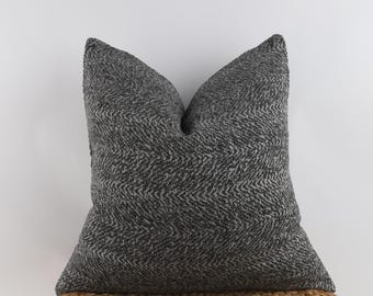 May include: A square decorative pillow with a dark gray and white herringbone pattern. The pillow is placed on a woven, light brown surface, and the background is a plain white.