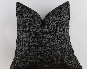 May include: A square decorative pillow with a textured black and gray pattern. The pillow has a soft, inviting appearance, perfect for adding a touch of style to any living space. The pillow is resting on a woven surface.